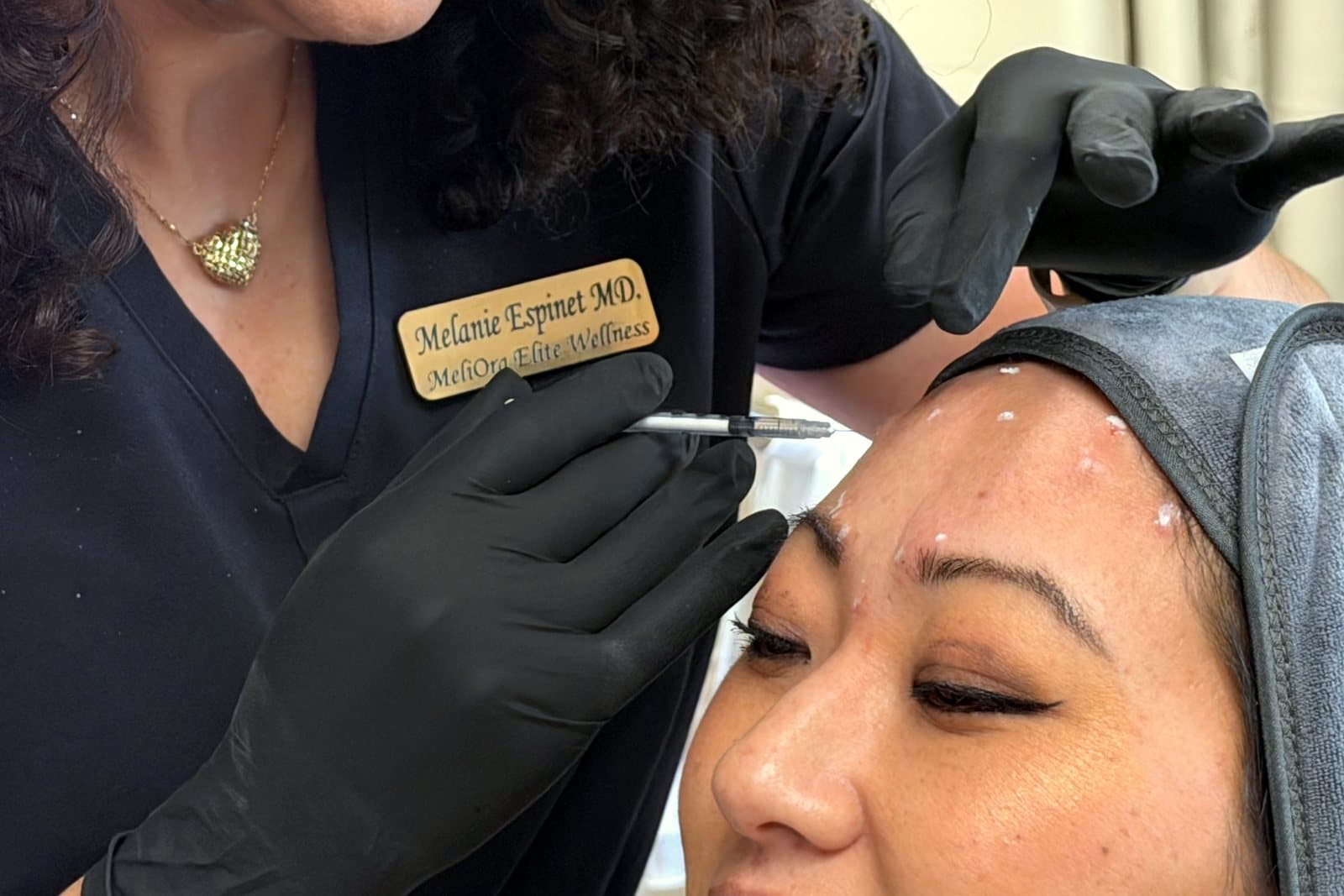 Black-gloved hands injecting a patient’s forehead below a name badge that reads Melanie Espinet MD