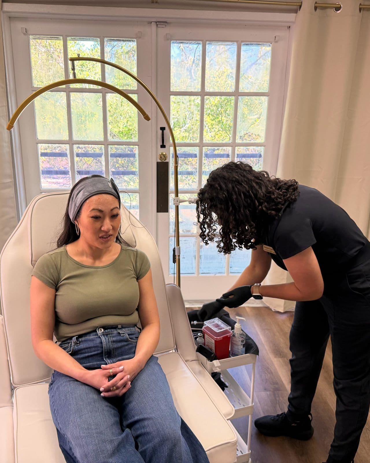 A patient in an olive T-shirt and gray headband sits in a treatment chair while Dr. Espinet prepares supplies
