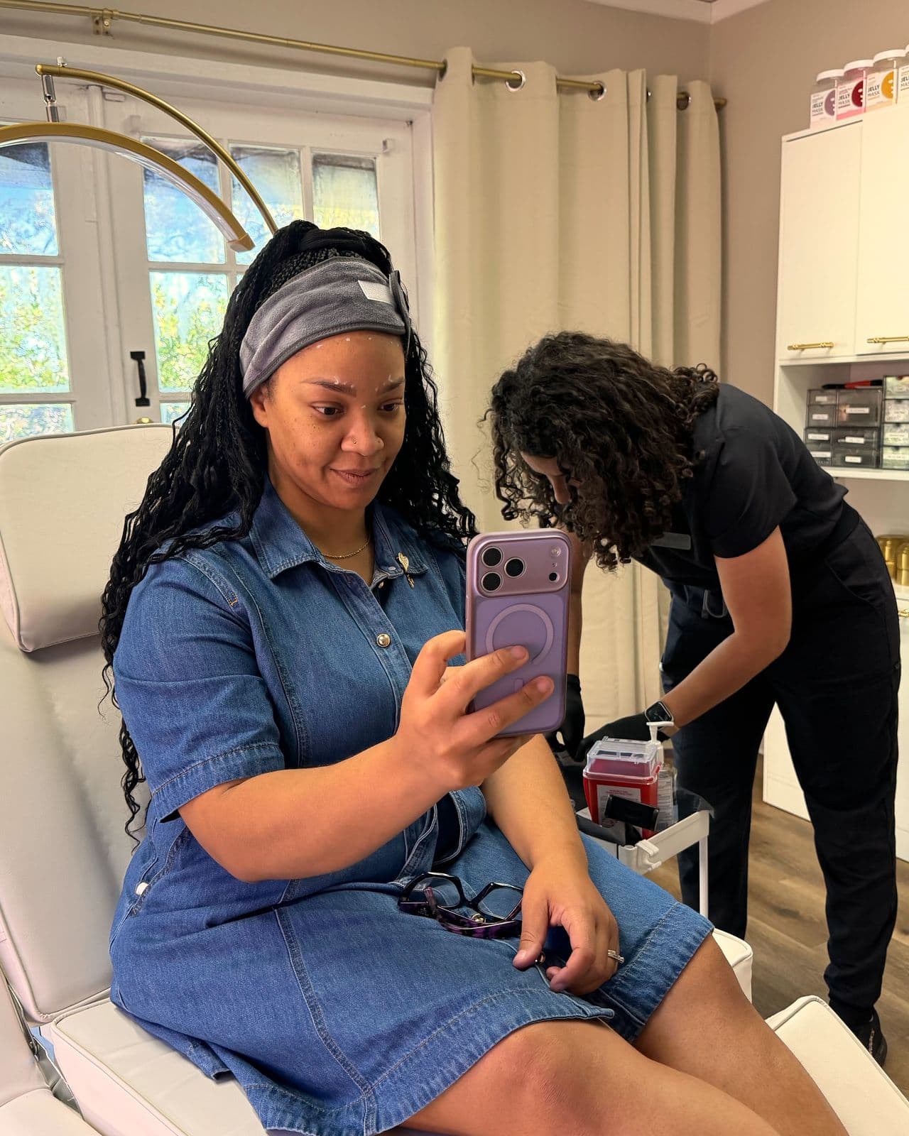 A patient in a denim dress and gray headband takes a selfie while Dr. Espinet prepares supplies behind her