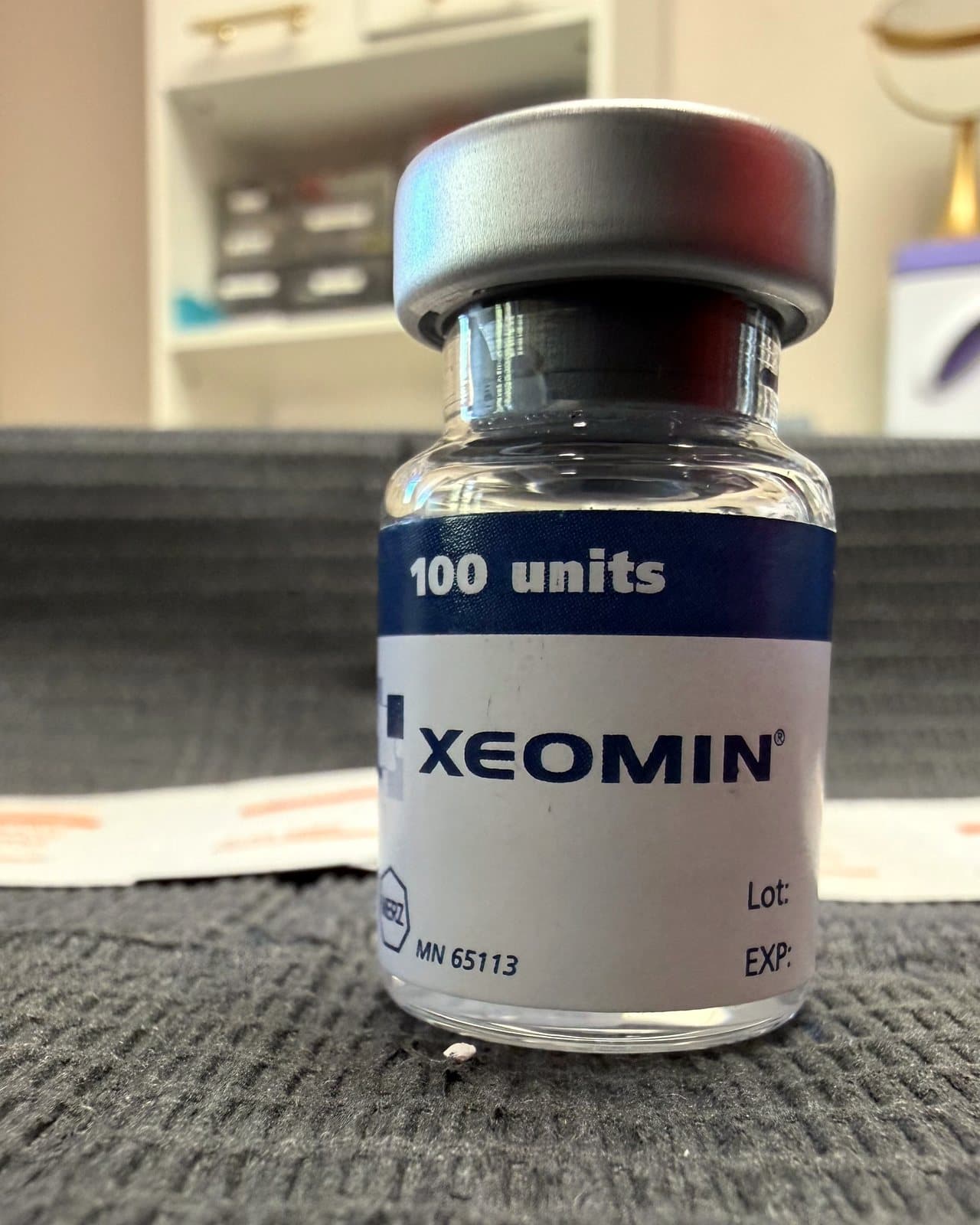 An upright 100-unit vial of Xeomin on a dark towel