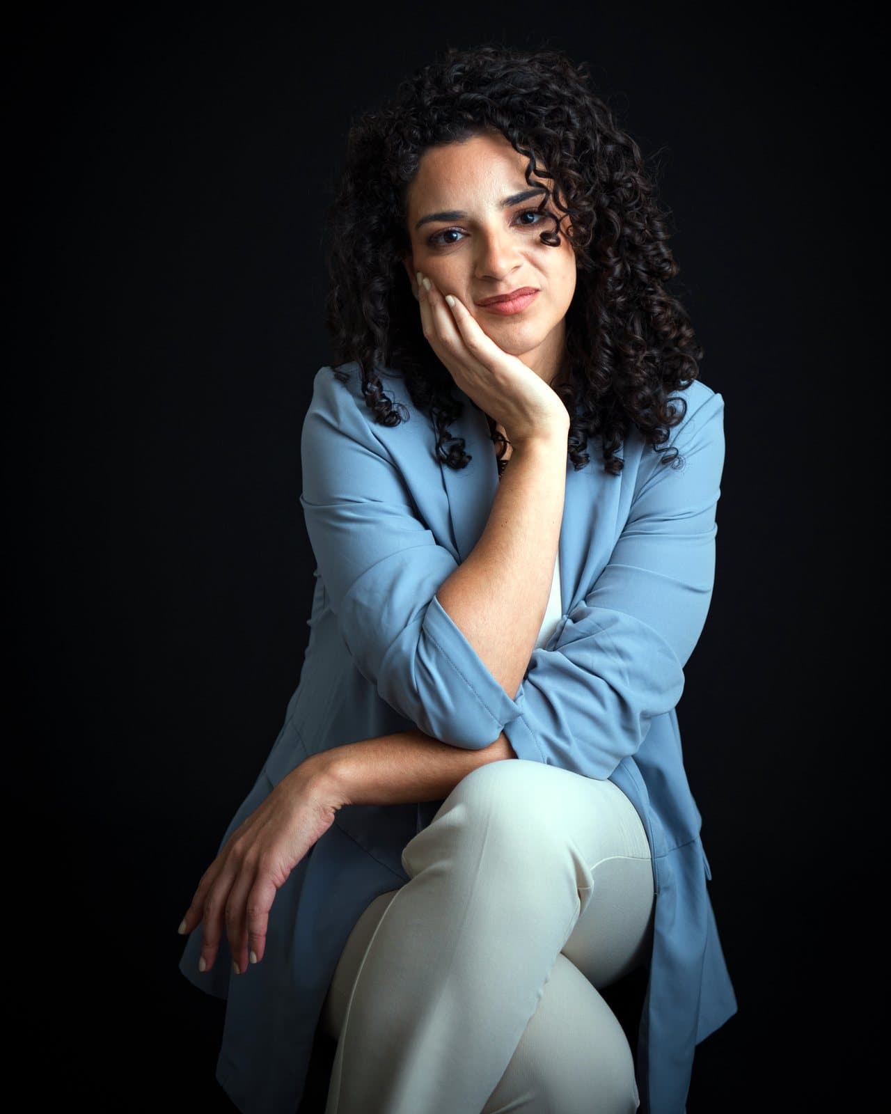 Dr. Melanie Espinet in a light blue blazer, seated with her chin resting on her hand, against a black backdrop
