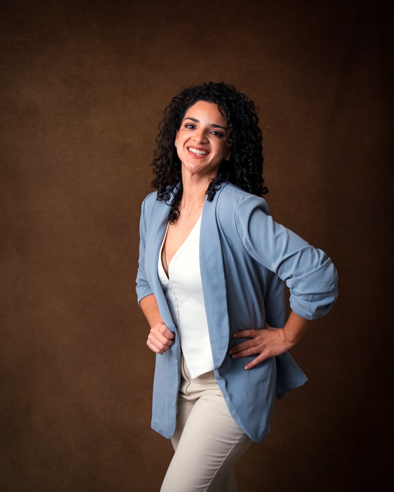 Dr. Melanie Espinet smiling in a light blue blazer with her hands on her hips, against a brown backdrop