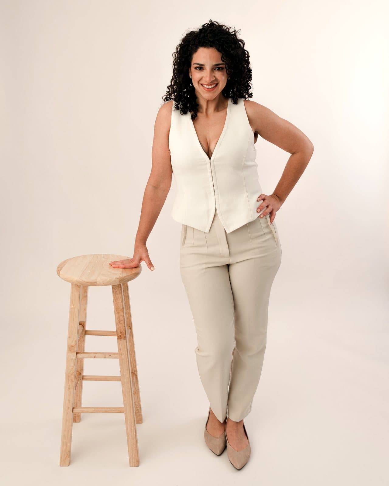 Dr. Melanie Espinet in a cream vest and beige trousers, standing with one hand on a wooden stool