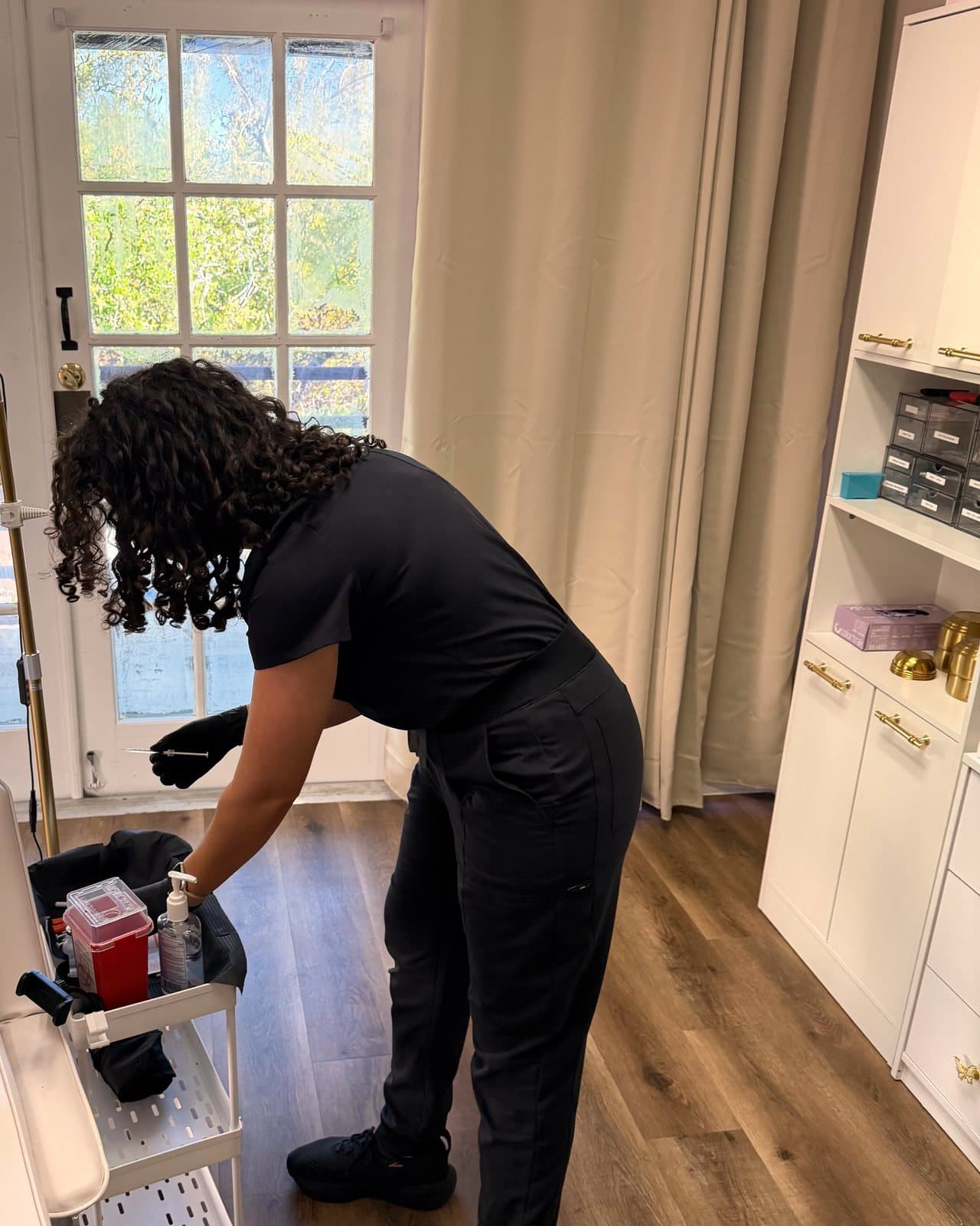 Dr. Espinet in black scrubs and gloves preparing supplies at a rolling cart with a red sharps container