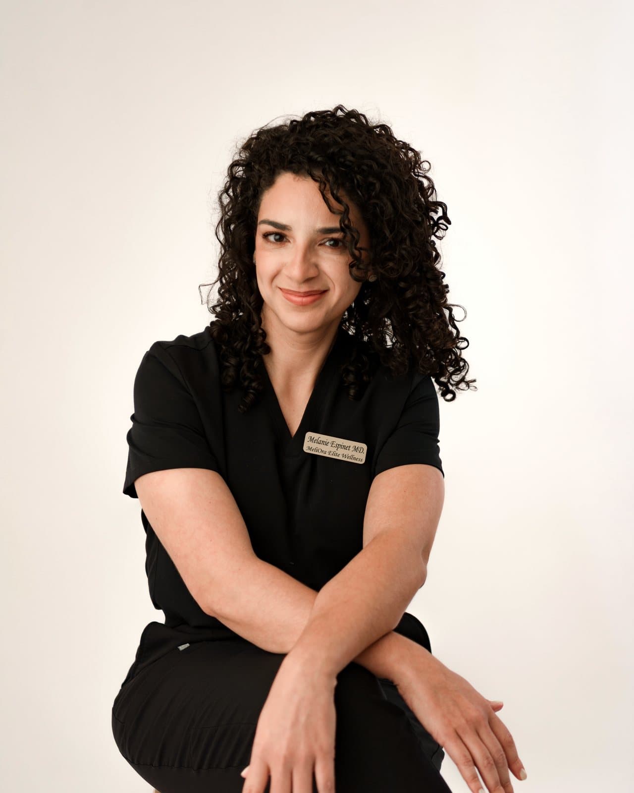 Dr. Melanie Espinet smiling in black scrubs, seated with her arms resting on her knee