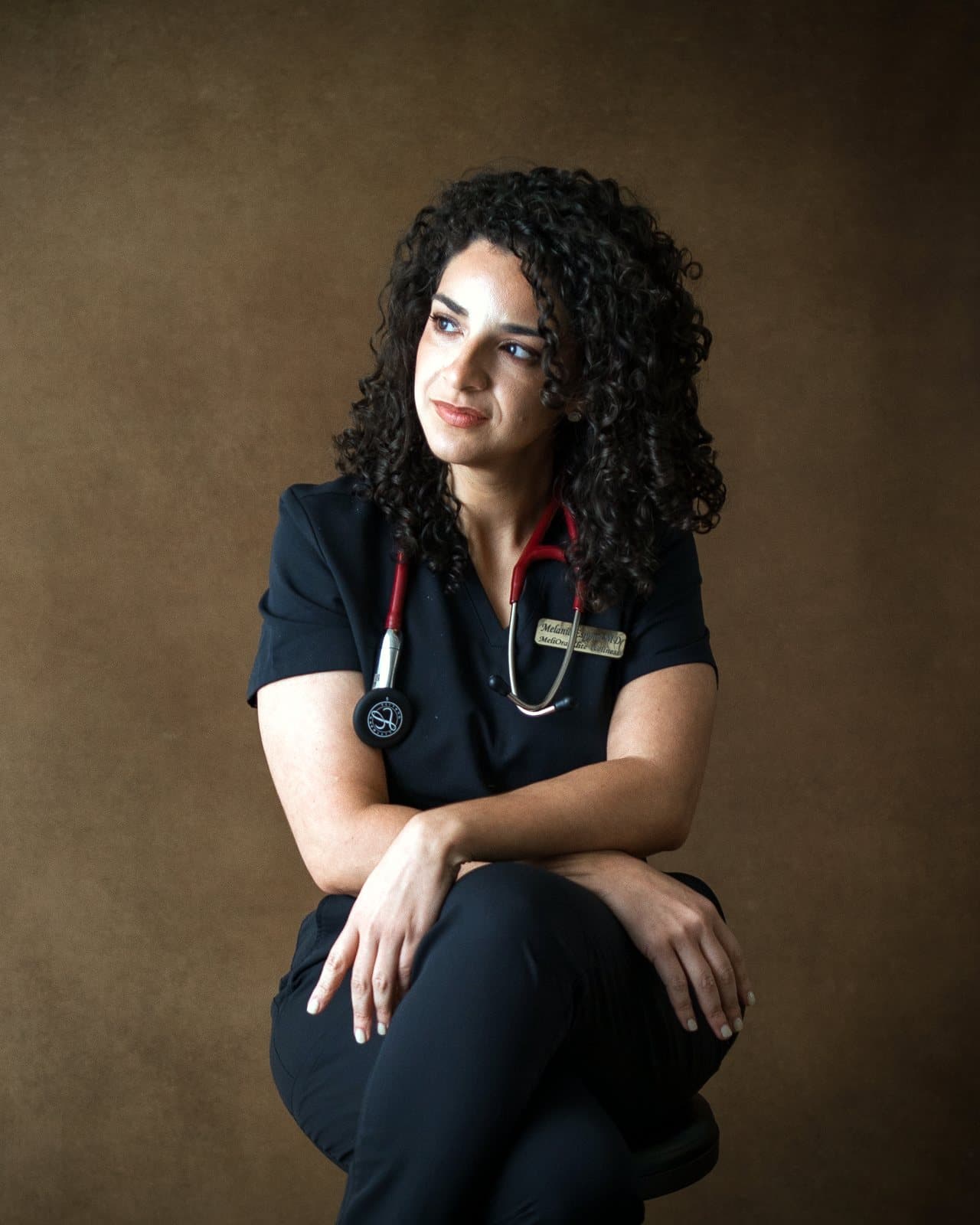 Dr. Melanie Espinet in black scrubs and a stethoscope, seated against a brown backdrop and looking to the side