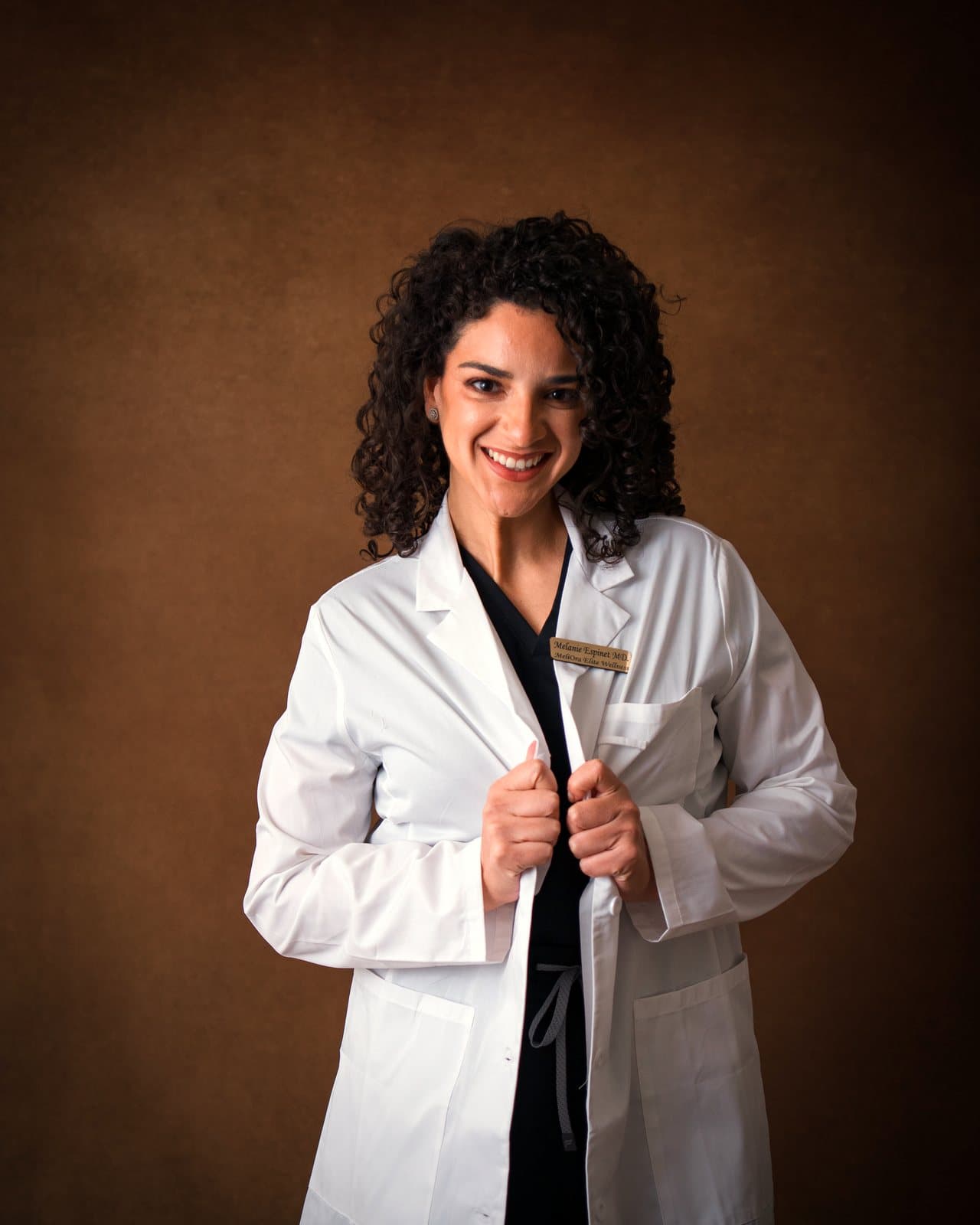 Dr. Melanie Espinet smiling in a white coat over black scrubs, holding the lapels, against a brown backdrop