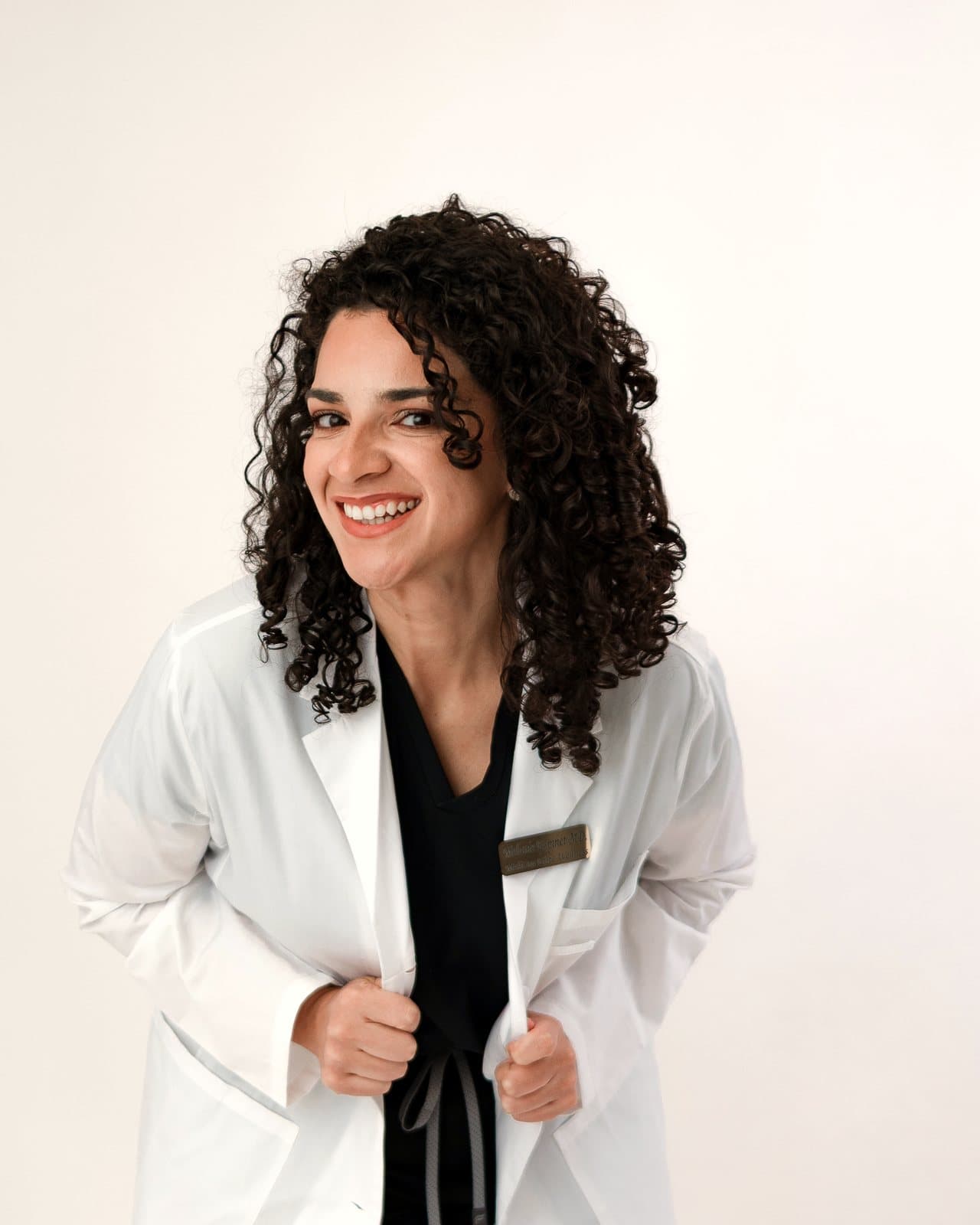 Dr. Melanie Espinet smiling broadly in a white coat over black scrubs, holding the lapels